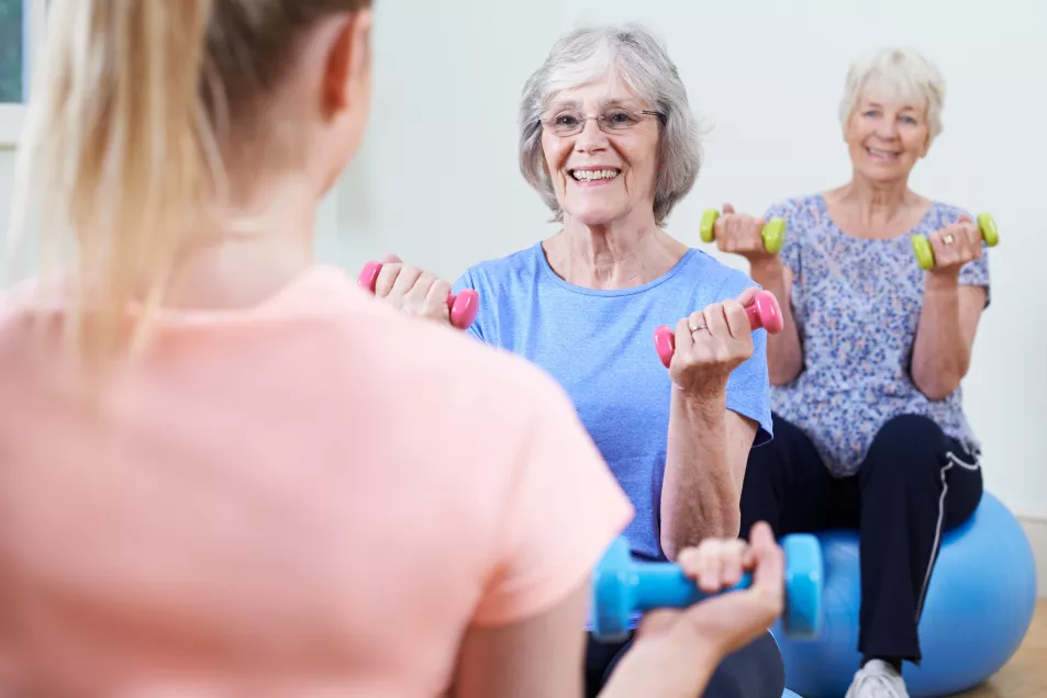 Two older women sitting on exercise balls lifting weights facing the instructor in a gym